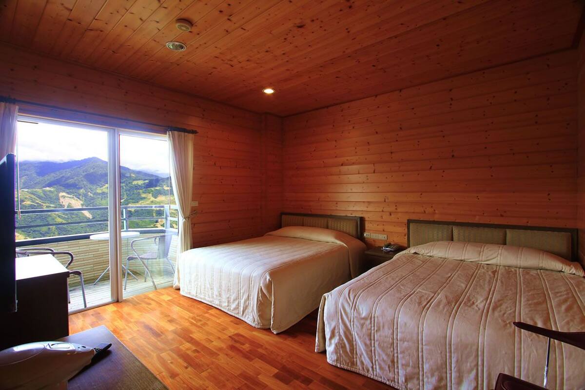 A wooden cabin room with two beds, a desk, and a balcony with a mountain view.