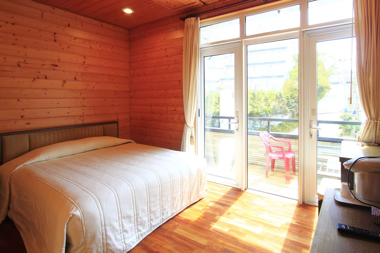 A wooden room with a bed, a sliding glass door to an outdoor area, and a table with a kettle.