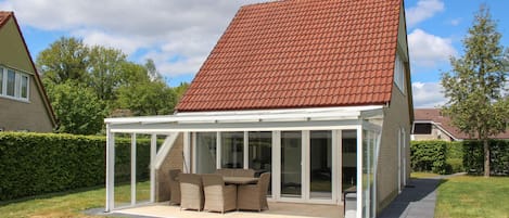 A house with a covered patio area featuring a table and chairs, surrounded by a hedge and a lawn.