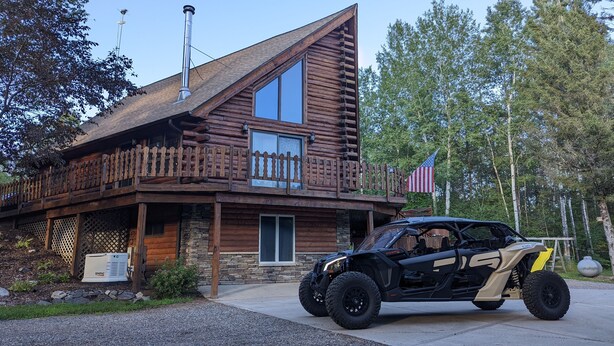 Une cabane en rondins dotée d’un balcon, avec des fondations en pierre, et un véhicule tout-terrain noir garé devant.