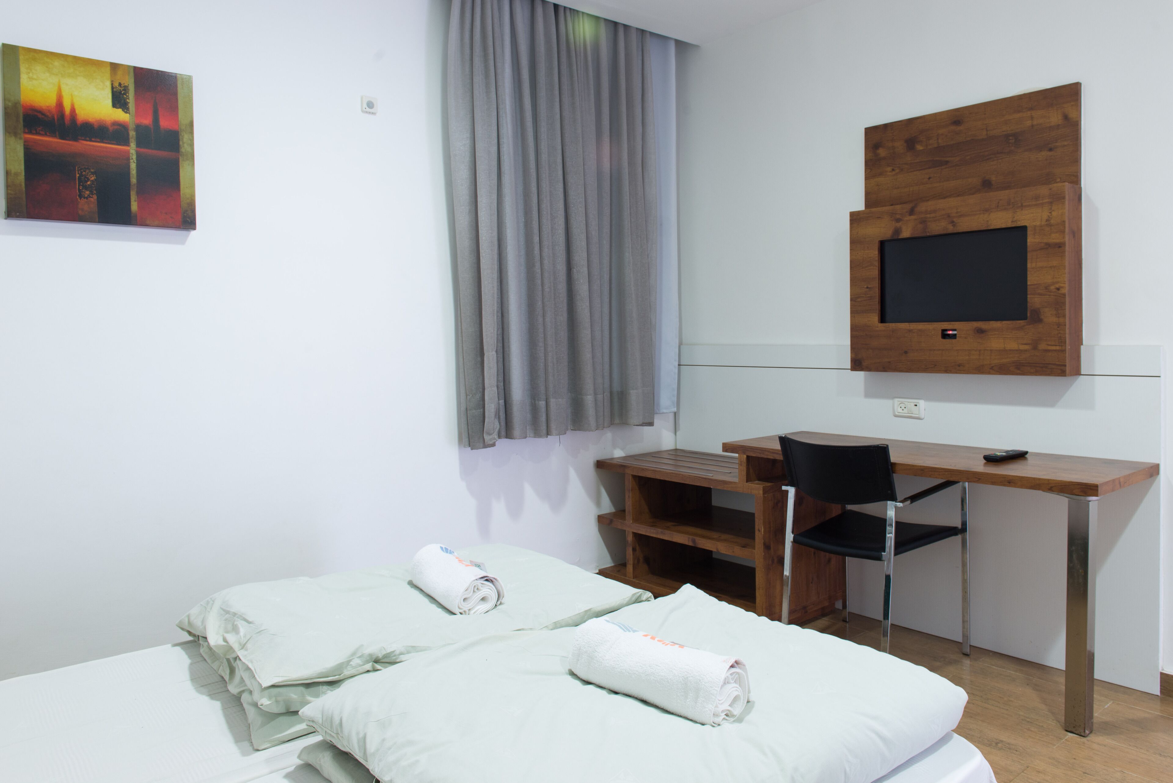 A room with two beds, a desk, a chair, a TV mounted on the wall, and a painting on the wall.