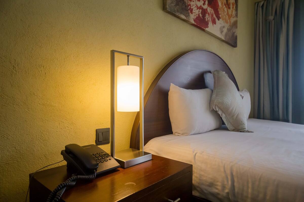 A hotel room with a bed, a bedside table with a phone, a lamp, and a wall-mounted artwork.