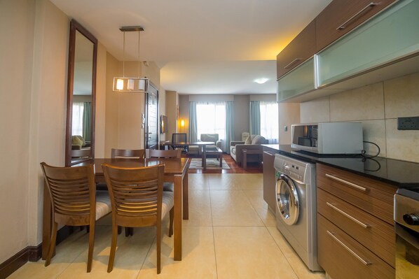 Cocina moderna con horno empotrado, microondas y lavadora. Cuenta con un comedor con mesa y sillas, además de una sala de estar con sofá y televisor.