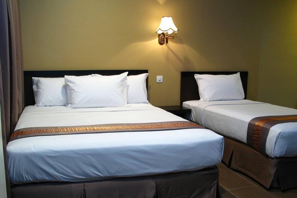 A hotel room with two beds, each with white linens and a brown accent. There is a wall-mounted lamp on the left side of the room.