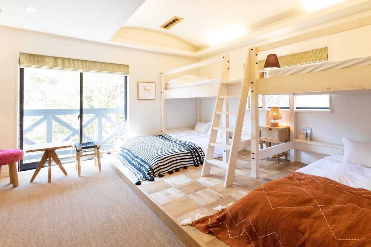 A cozy room with bunk beds, a desk with a lamp, and a view of greenery outside.