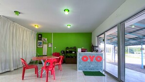 A room with a green wall, a reception desk with 'OYO' branding, a dining area with red chairs, and a large window.