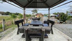 A covered outdoor seating area with a table and chairs, surrounded by greenery and buildings in the background.