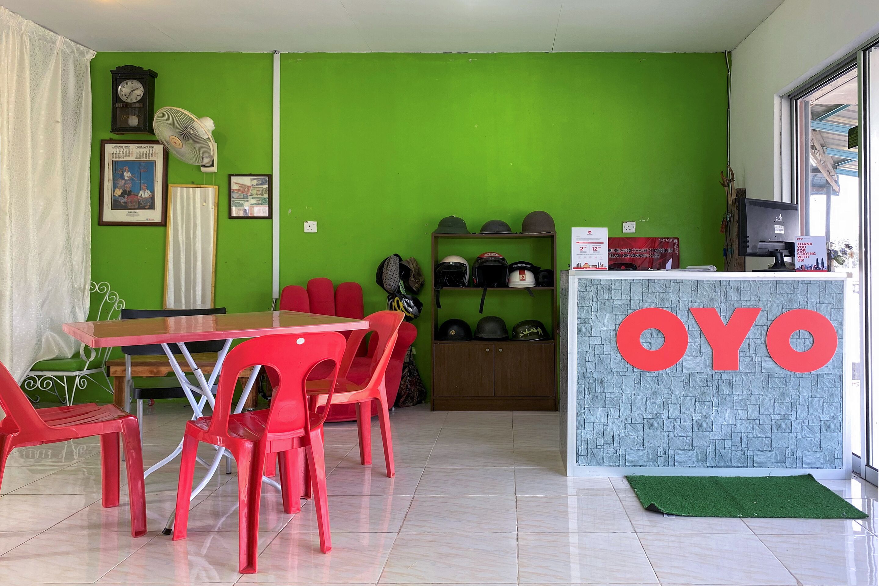 A room with a green wall, a wooden table with red chairs, a fan, and a shelf with helmets.