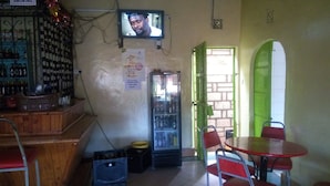 Un pequeño bar con televisor montado en la pared, un refrigerador y algunas sillas y mesas.