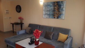 Un salón con un sofá gris, una mesita de centro blanca y un jarrón rojo. Hay un cuadro en la pared y una pequeña mesa con un jarrón de flores.