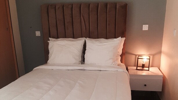 Una cama bien hecha con cabecera, una mesita de noche con lámpara y un reloj de pared.