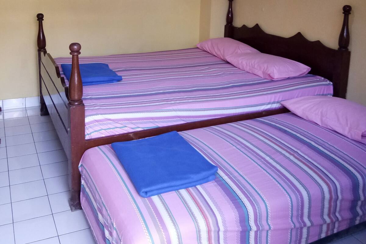 A room with two beds, a wooden headboard, a ceiling vent, and tiled flooring.