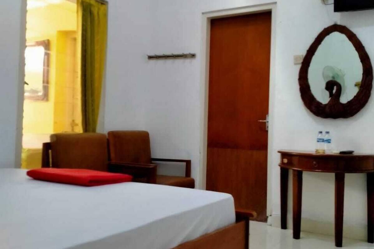 A hotel room with a bed, a chair, a table with a mirror, and an air conditioning unit on the wall.