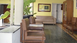 A living room with a beige sofa, wooden chairs, a cabinet, and a large plant. - Prayogolama Guest House (Yogyakarta)