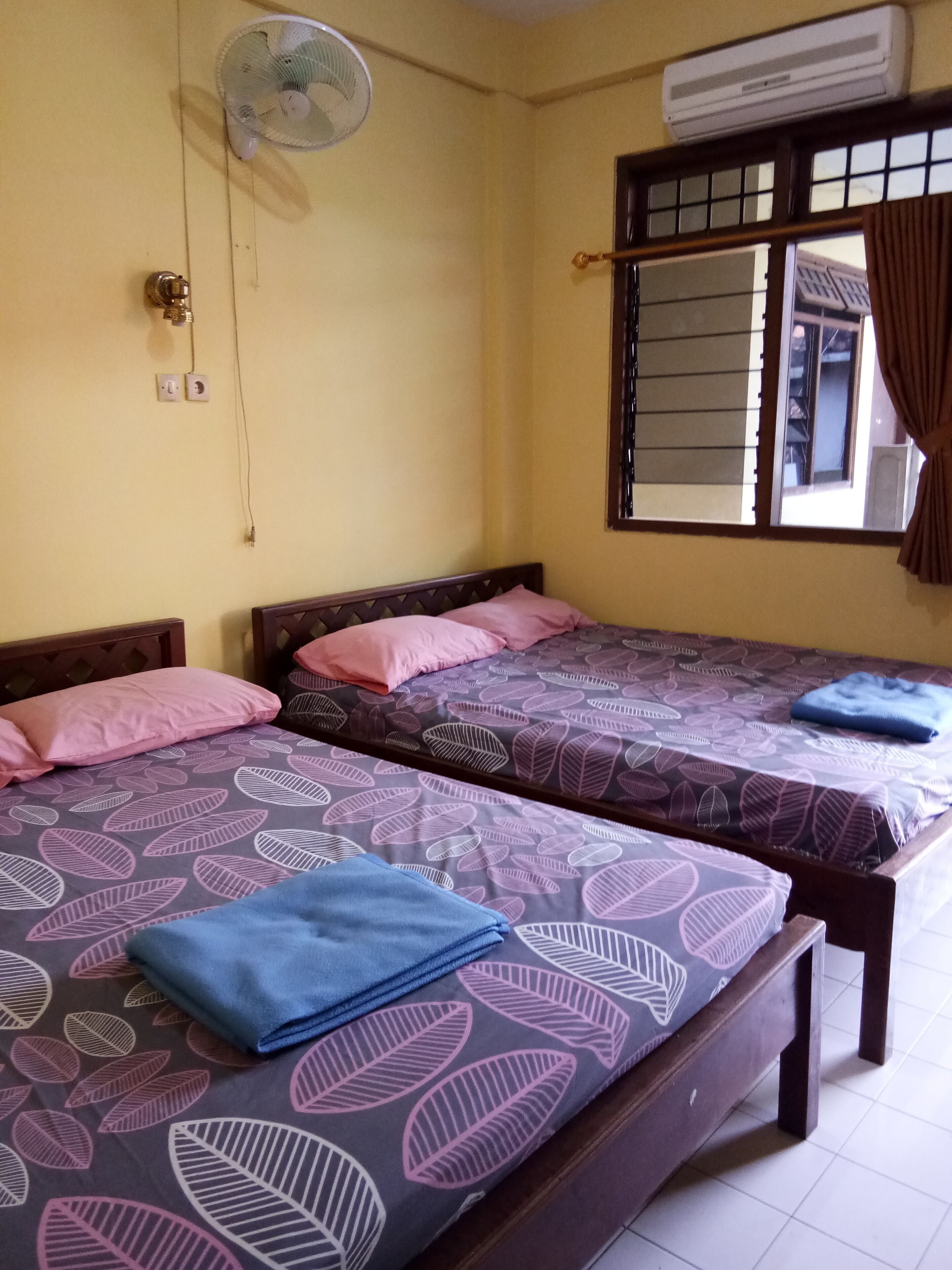 Una habitación con dos camas, cada una con colcha y almohadas estampadas, un ventilador de techo, un equipo de aire acondicionado y una ventana con cortinas.