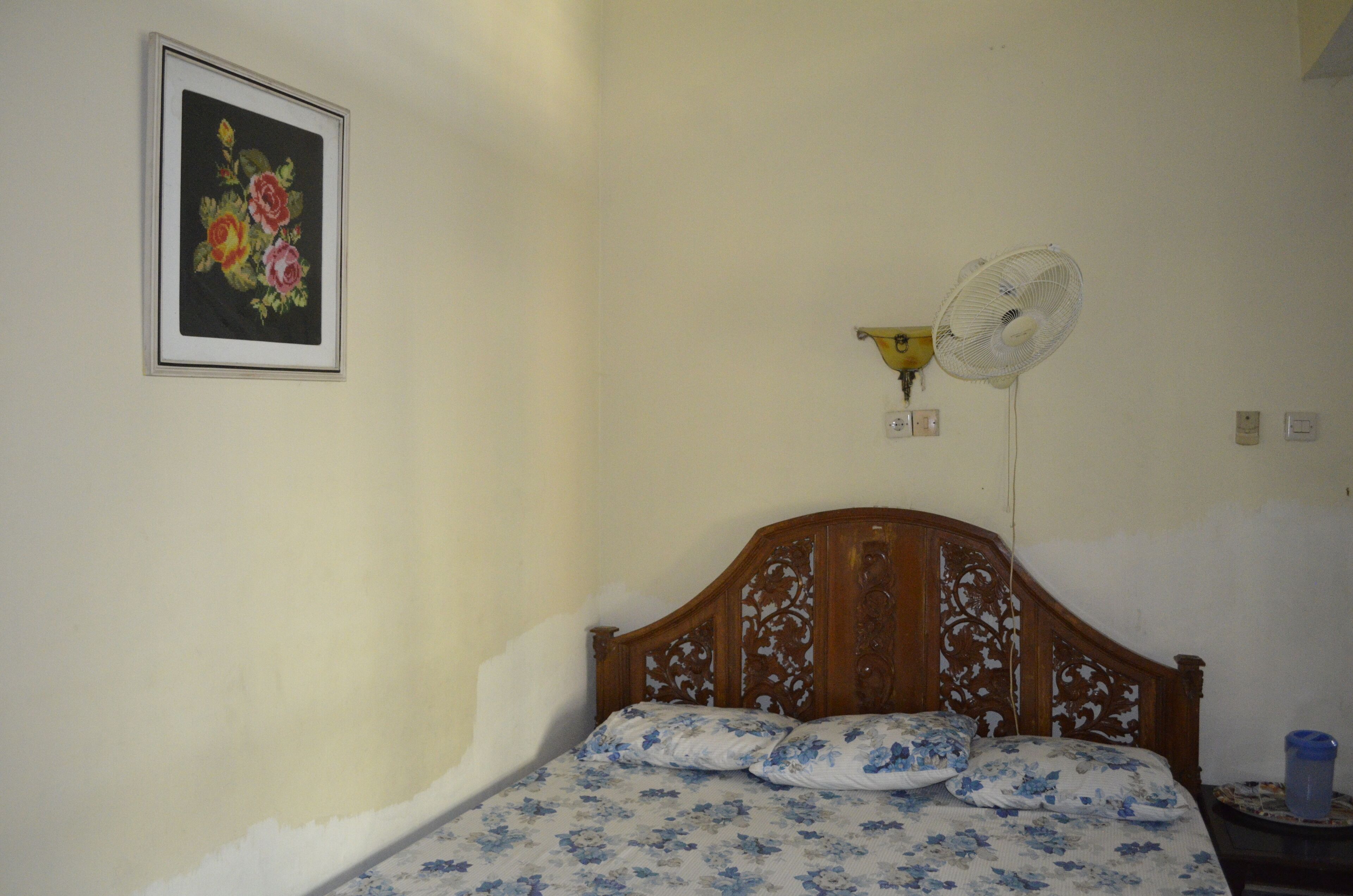 Un dormitorio con cabecera de madera, cama con ropa de cama floral, ventilador de pared y una obra de arte floral enmarcada.
