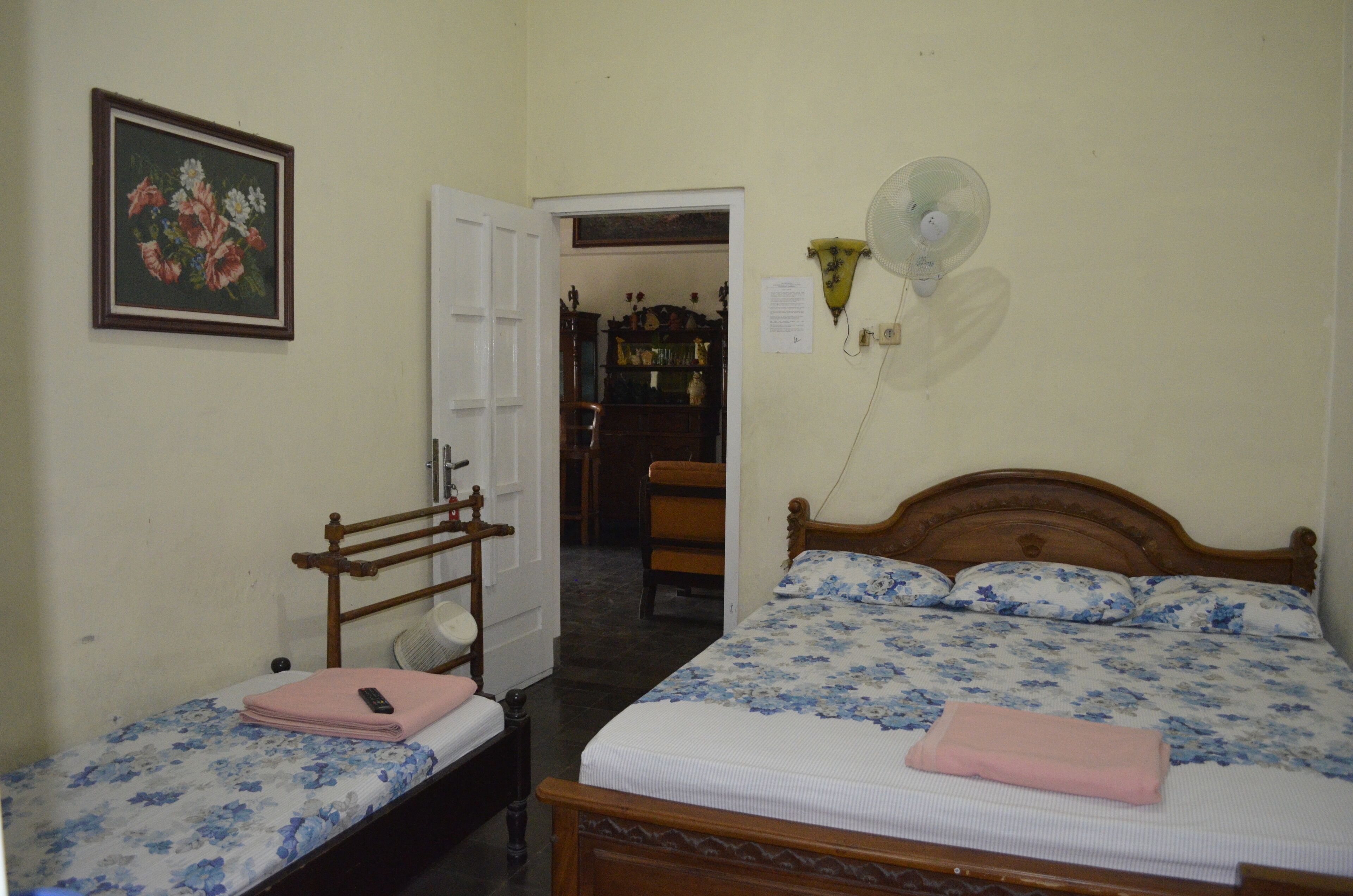 A bedroom with two beds, a wooden headboard, a bedside table, a chair, a fan, and a door leading to another room.