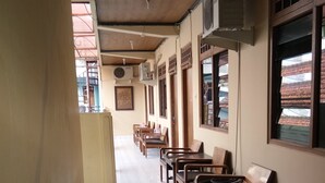 A corridor with wooden chairs and tables, air conditioning units, and windows with views of buildings. - Prayogolama Guest House (Yogyakarta)