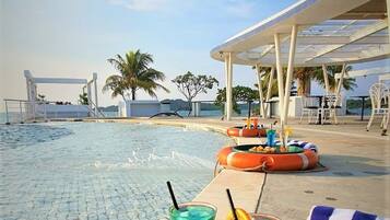 Escena junto a la piscina con una mesa preparada para una comida, incluyendo sándwiches y bebidas, y vista al mar y palmeras.