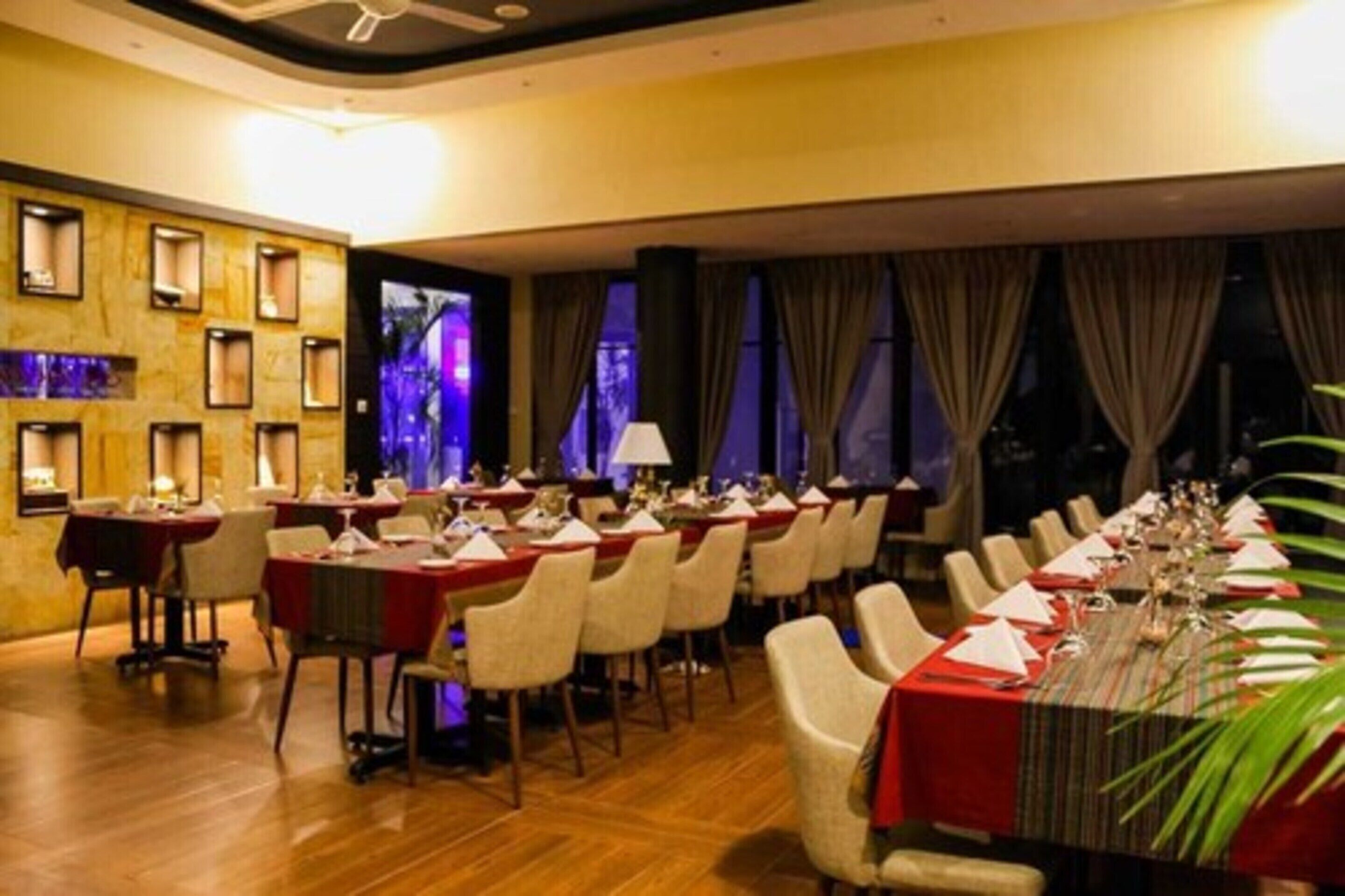 A restaurant with multiple tables set for dining, a wooden wall with framed pictures, and a view of the outdoors through large windows.
