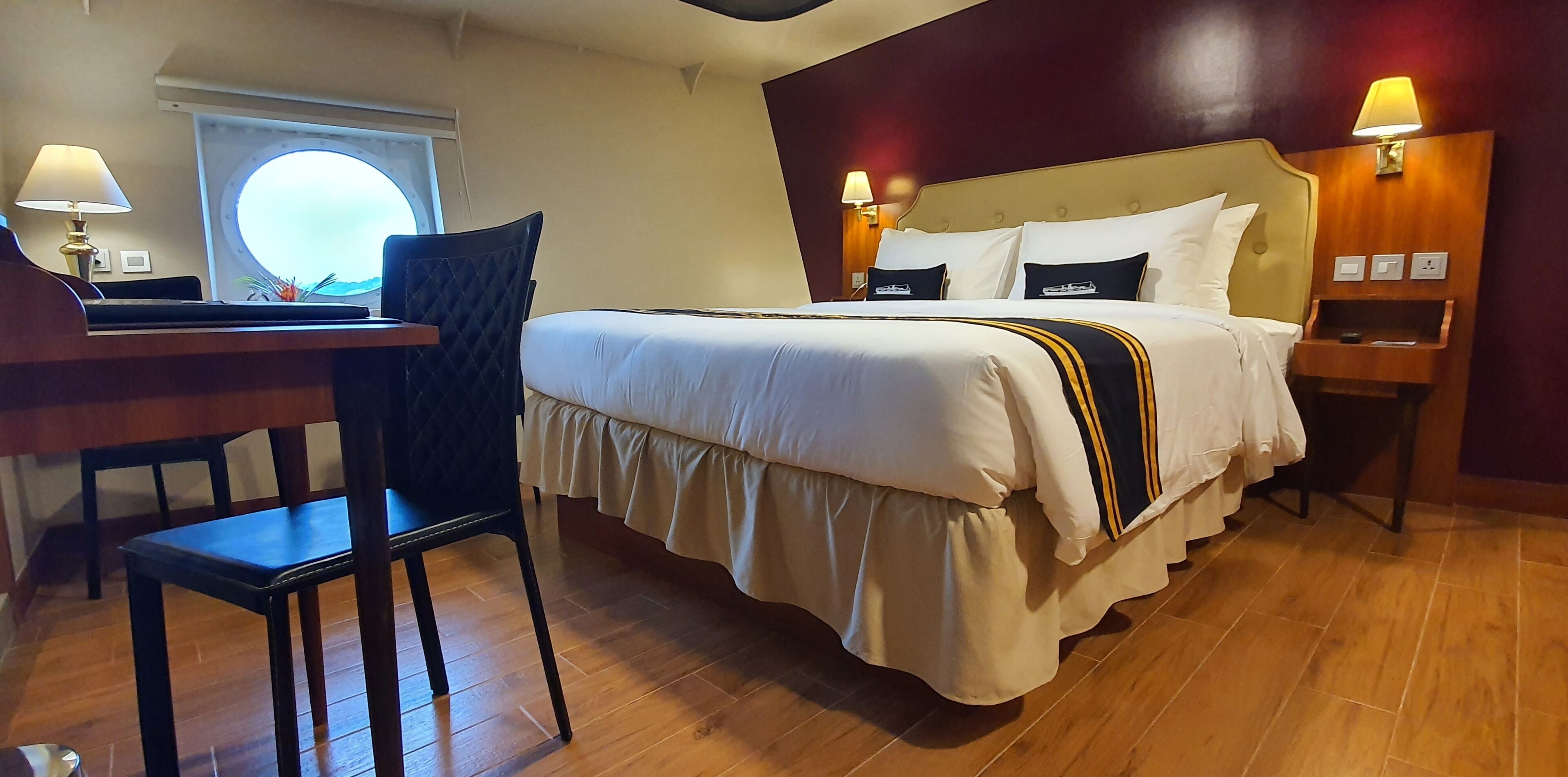 Una habitación de hotel con cama, escritorio, silla, ventana y mesita de noche con lámpara.