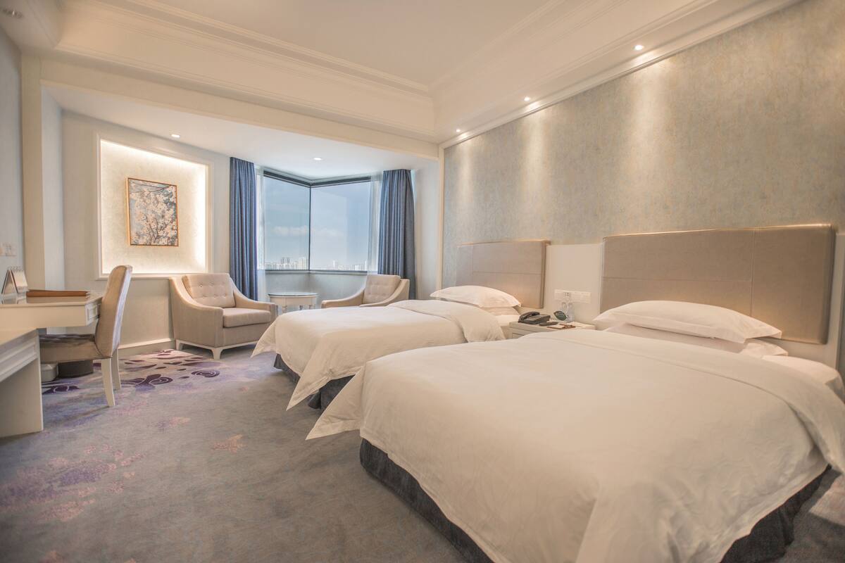 A hotel room with two beds, a desk, a chair, and a large window with a view.