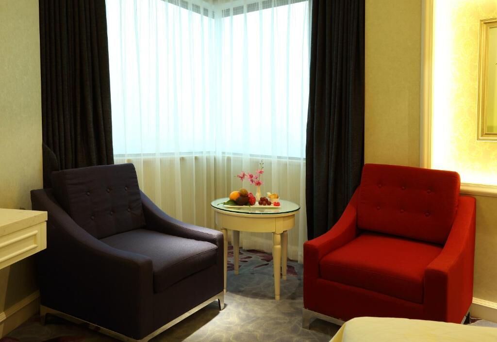 A hotel room with two armchairs, a small table with fruit and flowers, and a window with sheer curtains.