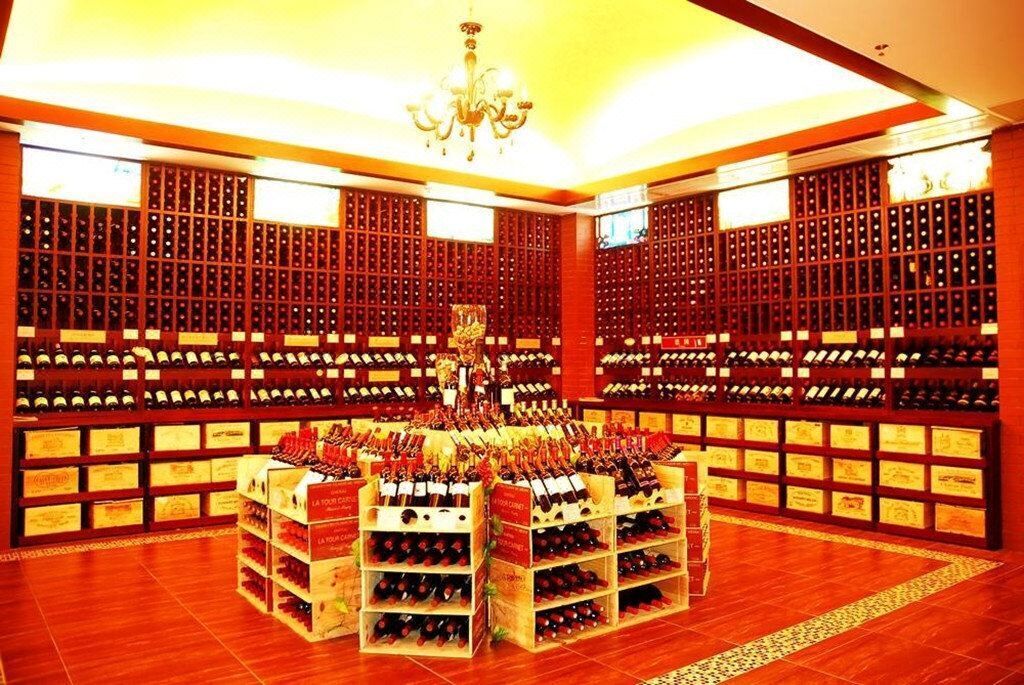 A wine cellar with wooden racks filled with bottles, a central display of wine boxes, and a chandelier.