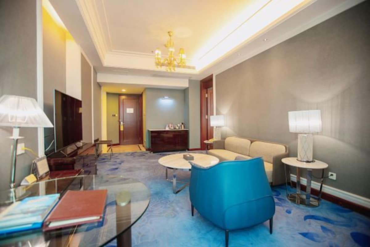 Una habitación de hotel moderna con una mesa de centro de vidrio, un sillón azul, un sofá beige y una pequeña mesa redonda con una lámpara.