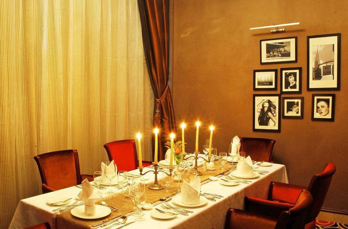 Comedor con una larga mesa dispuesta para una comida, cuadros en la pared y una cortina en la ventana.