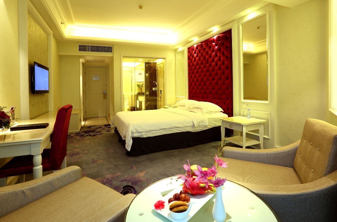 Una habitación de hotel con una cama, un escritorio con una silla, una mesa redonda con un jarrón de flores y un cabecero tapizado en rojo.