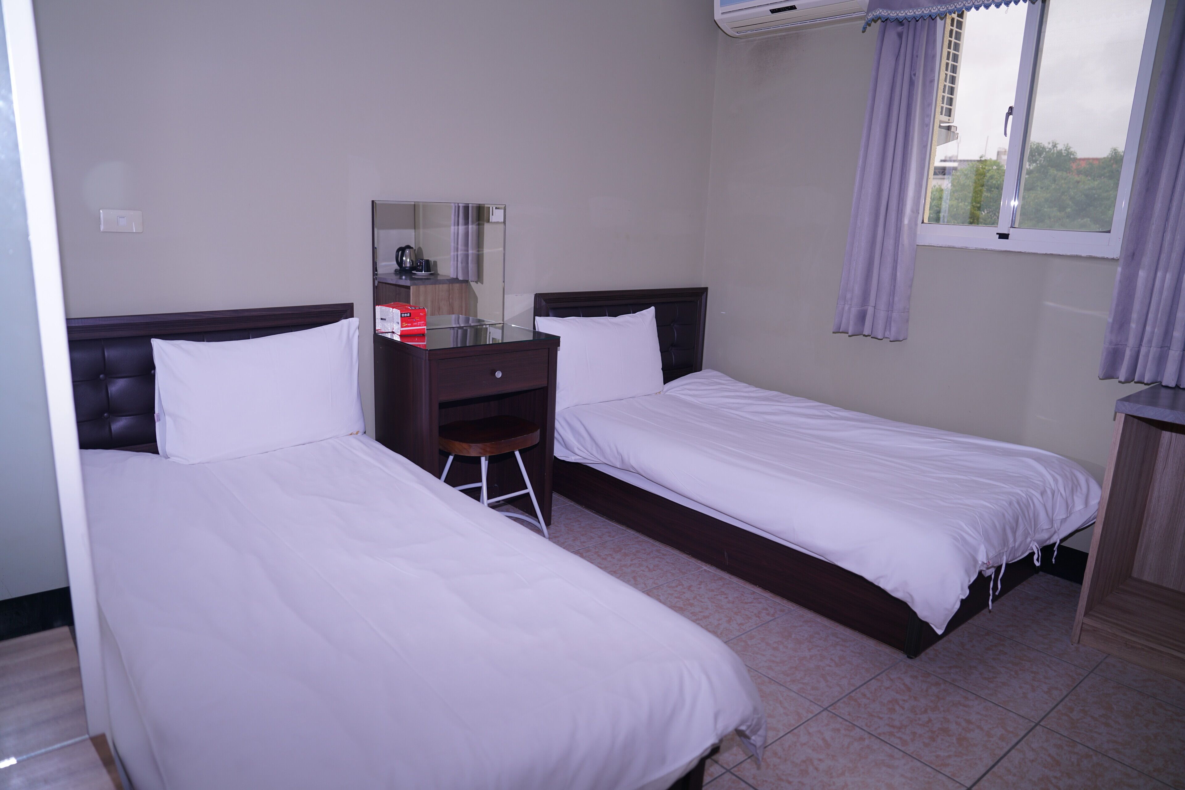 Habitación de hotel con dos camas individuales, un escritorio pequeño con silla, un espejo y una ventana con cortinas.