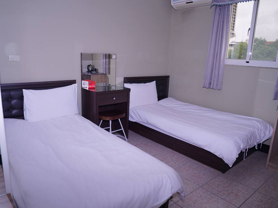 Habitación de hotel con dos camas individuales, un escritorio pequeño con silla, un espejo y una ventana con cortinas.