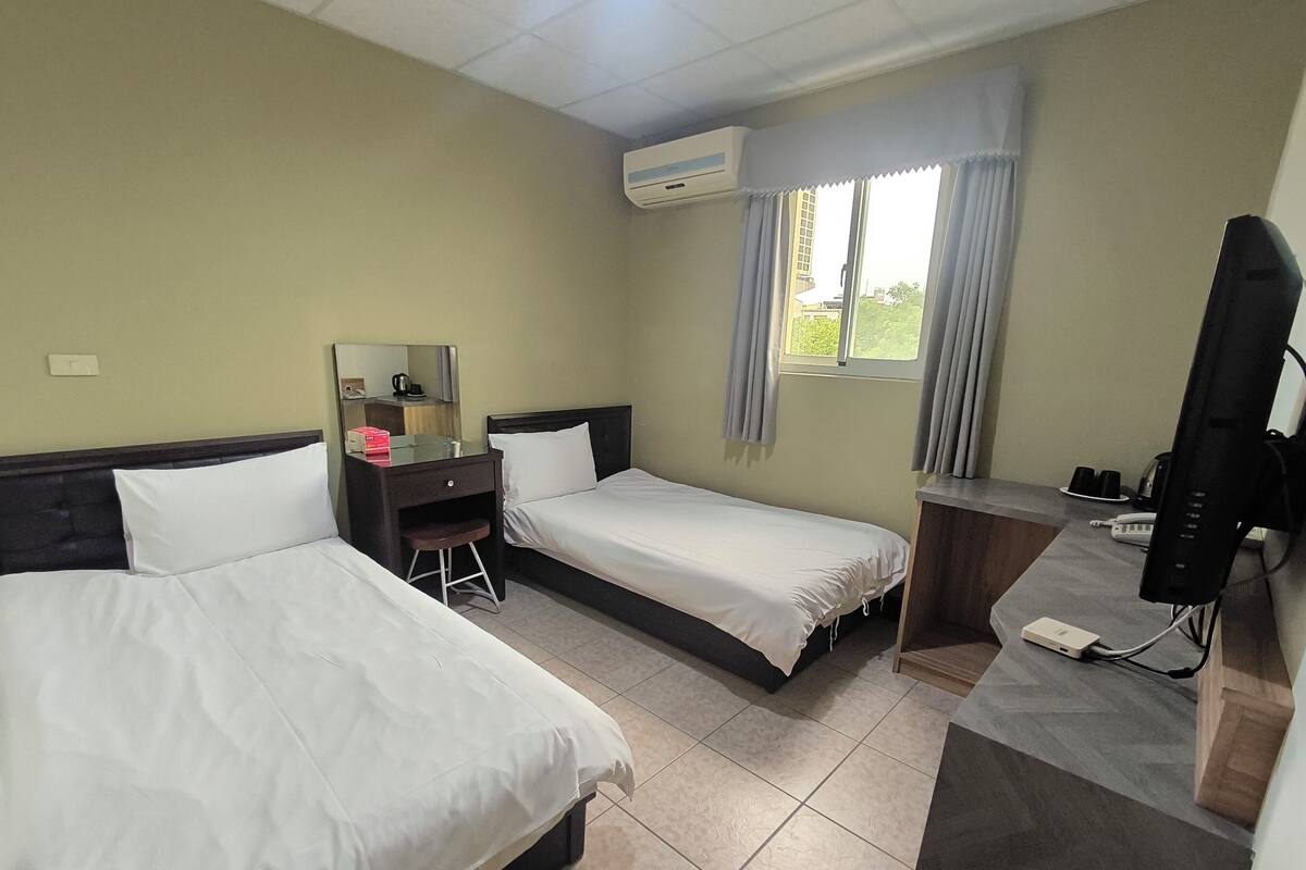 A hotel room with two beds, a desk, a chair, a TV, and an air conditioning unit.