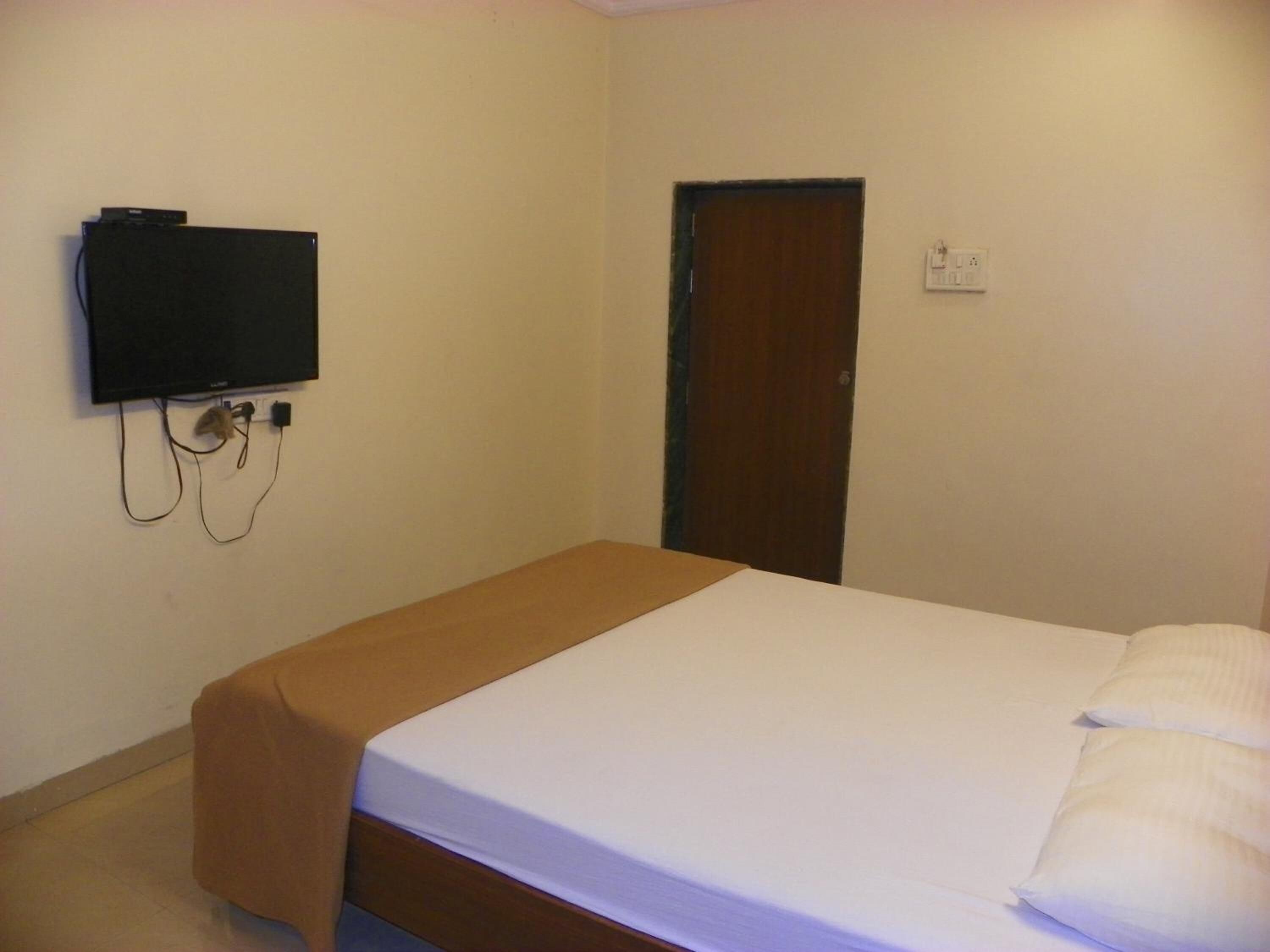 A single bed with a white mattress and a brown headboard, a flat-screen TV mounted on the wall, and a wooden door.