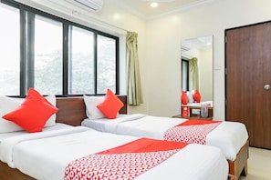 A hotel room with two beds, each with white bedding and red pillows. There is a large window with curtains, a wooden door, and a mirror on the wall. - Hotel Rajdeep (Pune)