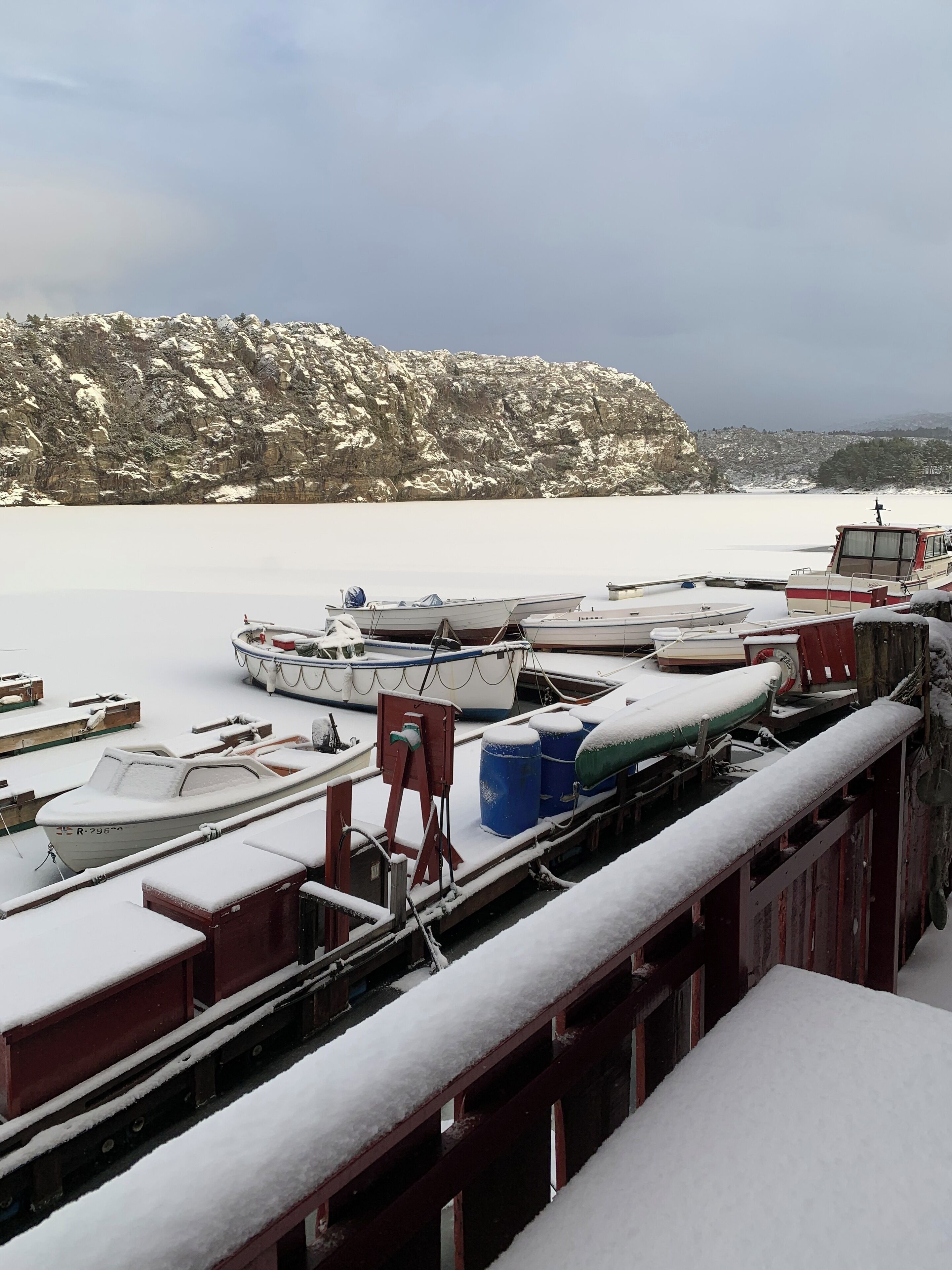 Un quai où sont amarrés plusieurs bateaux recouverts de neige, une falaise rocheuse en arrière-plan et un ciel dégagé.