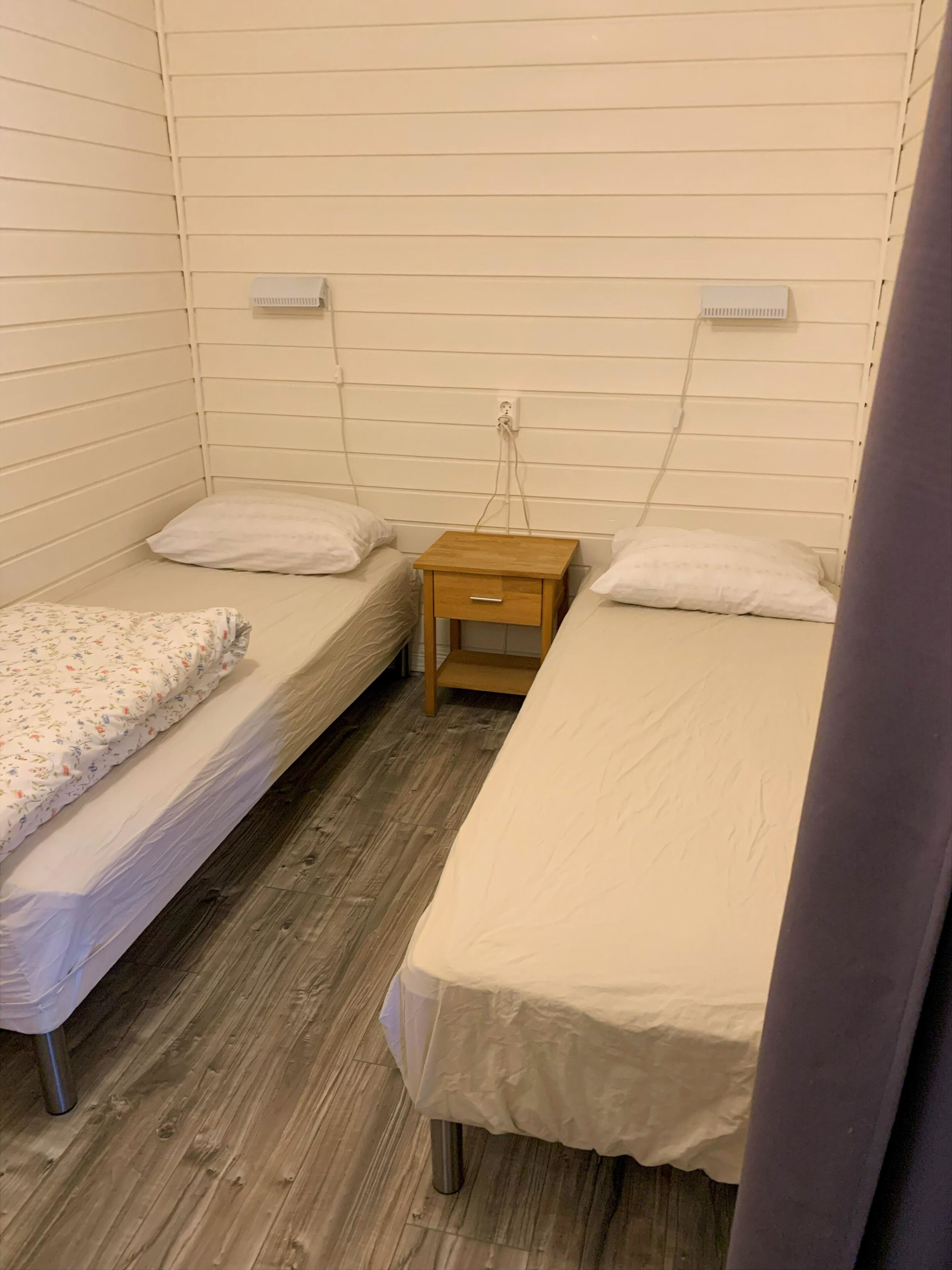 A small, single-room accommodation with two single beds, a wooden nightstand, and a wall-mounted heater.