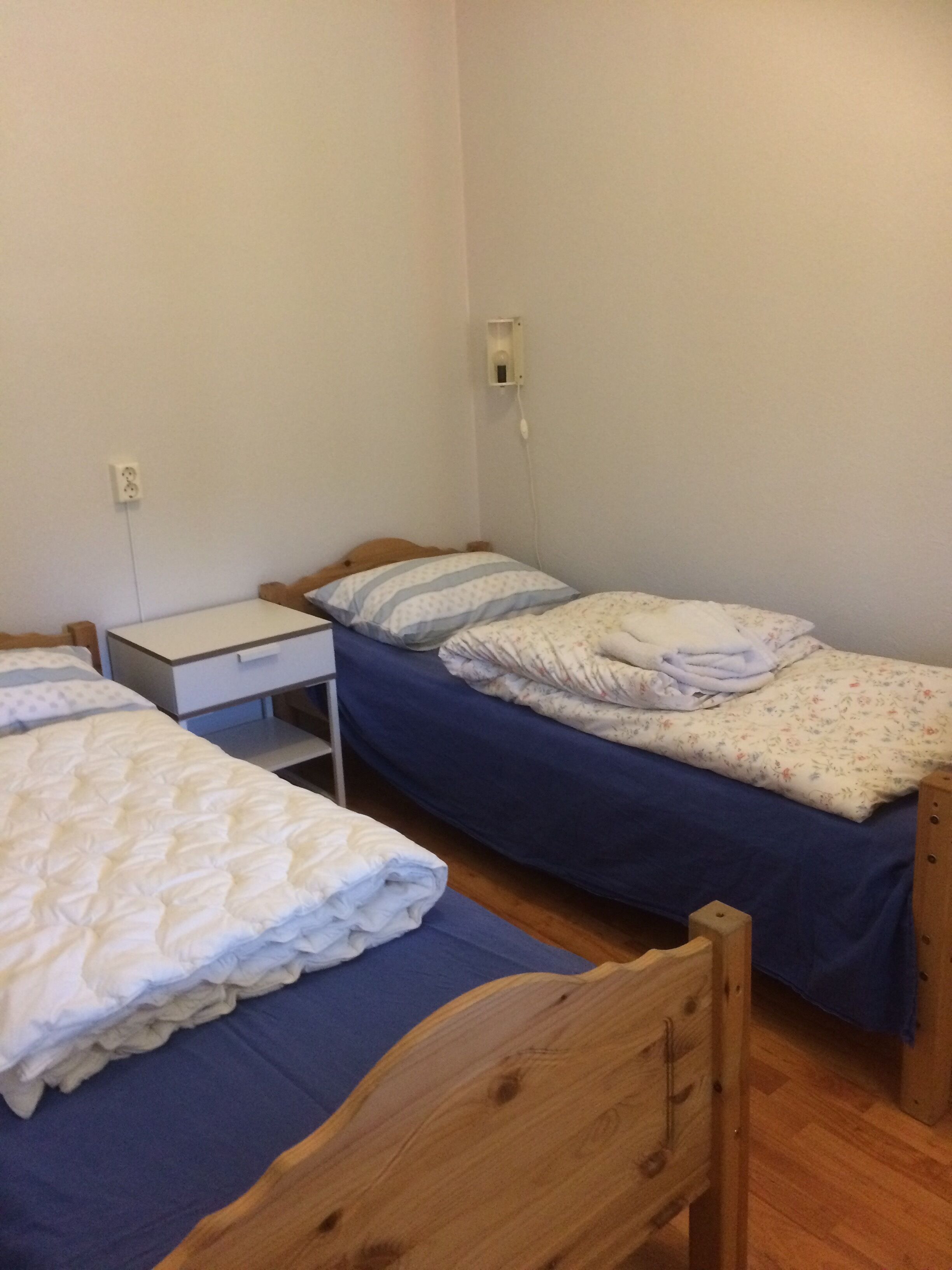 A small, simple room with two single beds, a wooden nightstand, and a wall socket.
