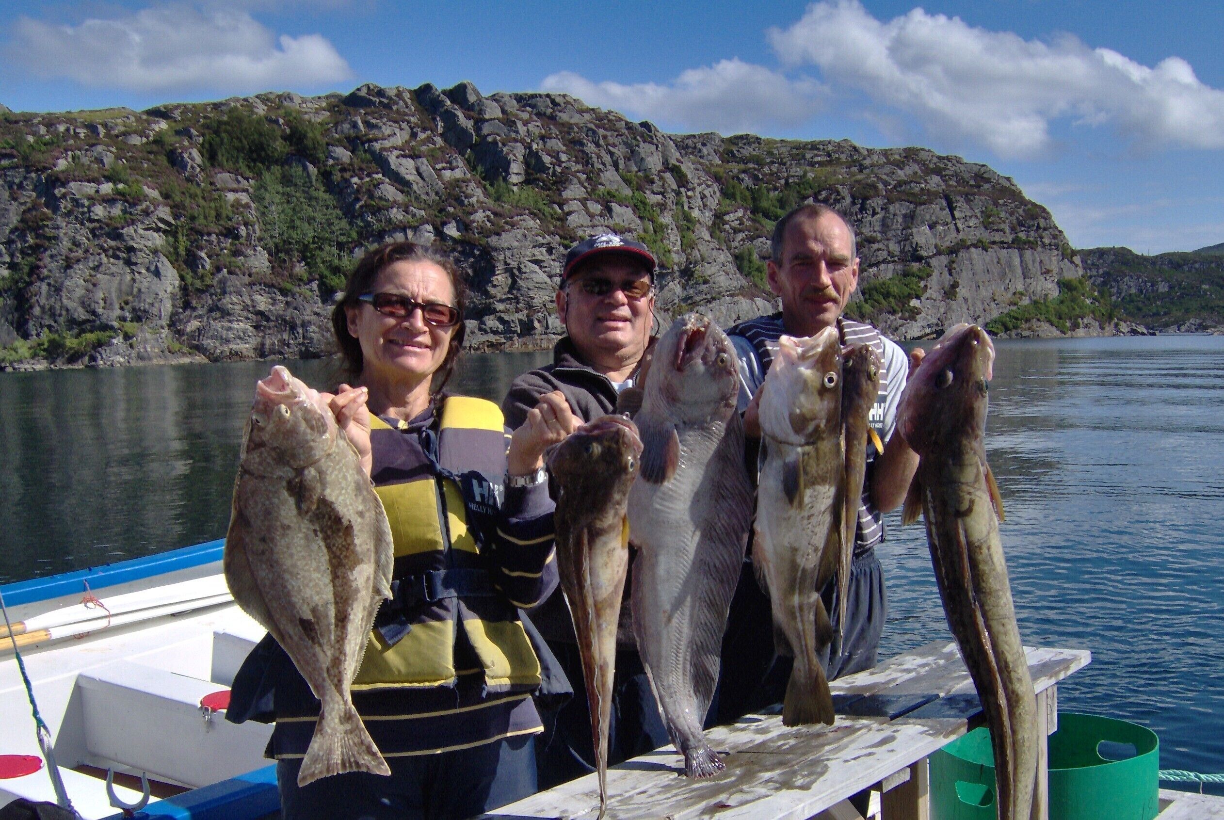 Three people holding fish on a boat with a rocky shoreline in the background.