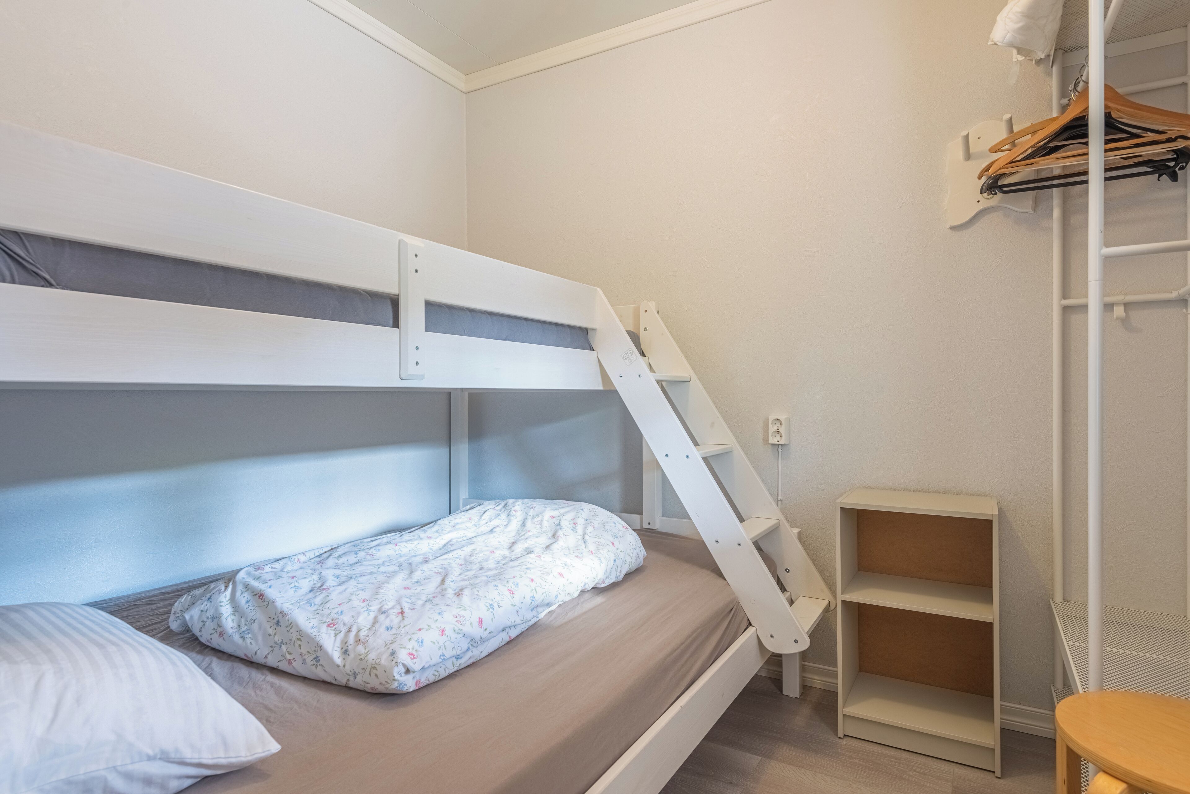 Un llit superposat amb escala, una taula de fusta petita i una estanteria en una habitació de parets blanques i terra de fusta.