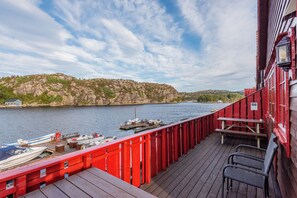 A wooden deck with red railings overlooking a calm body of water, boats docked, and a rocky hillside in the background.