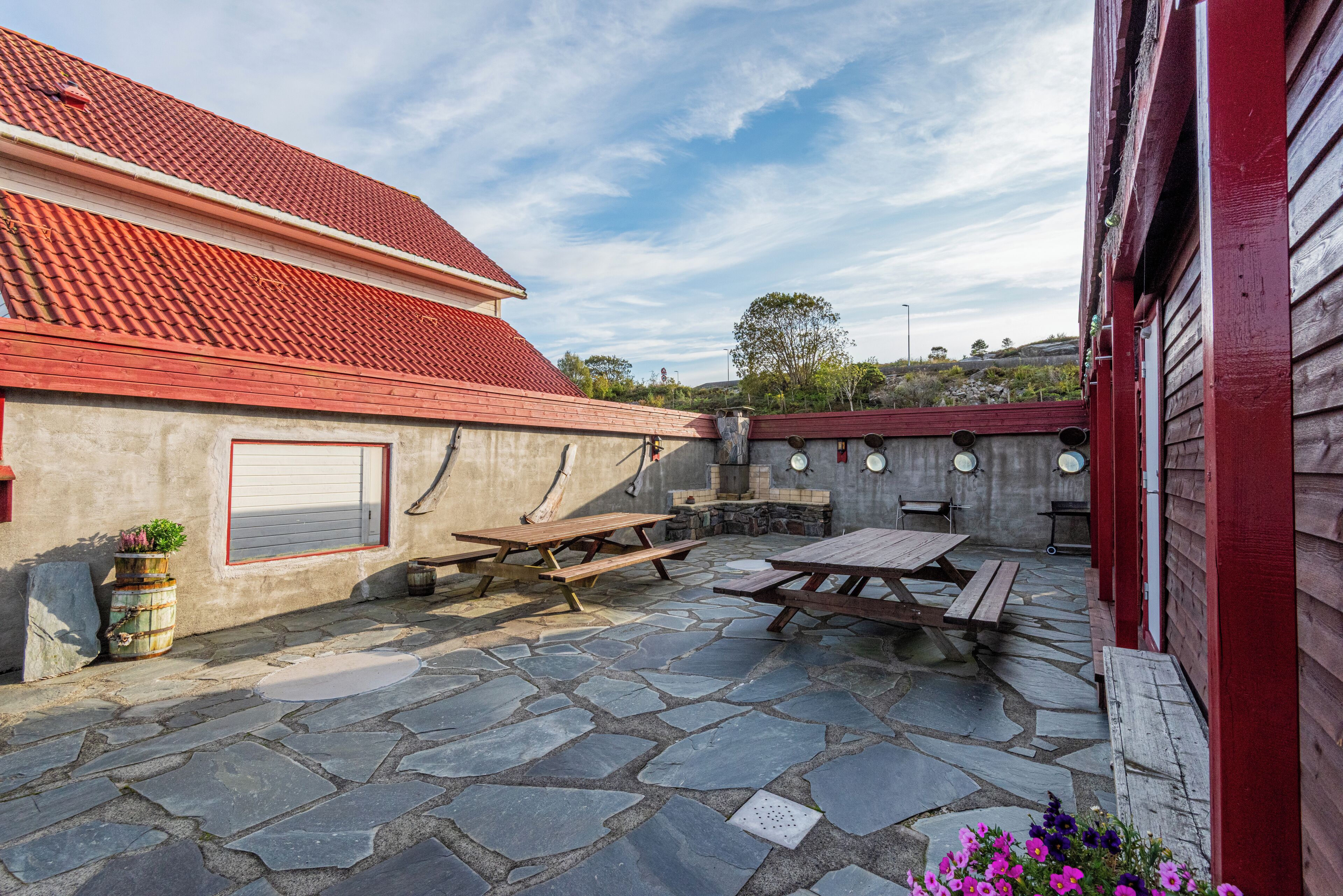 A patio area with stone flooring, picnic tables, and a red-roofed building.