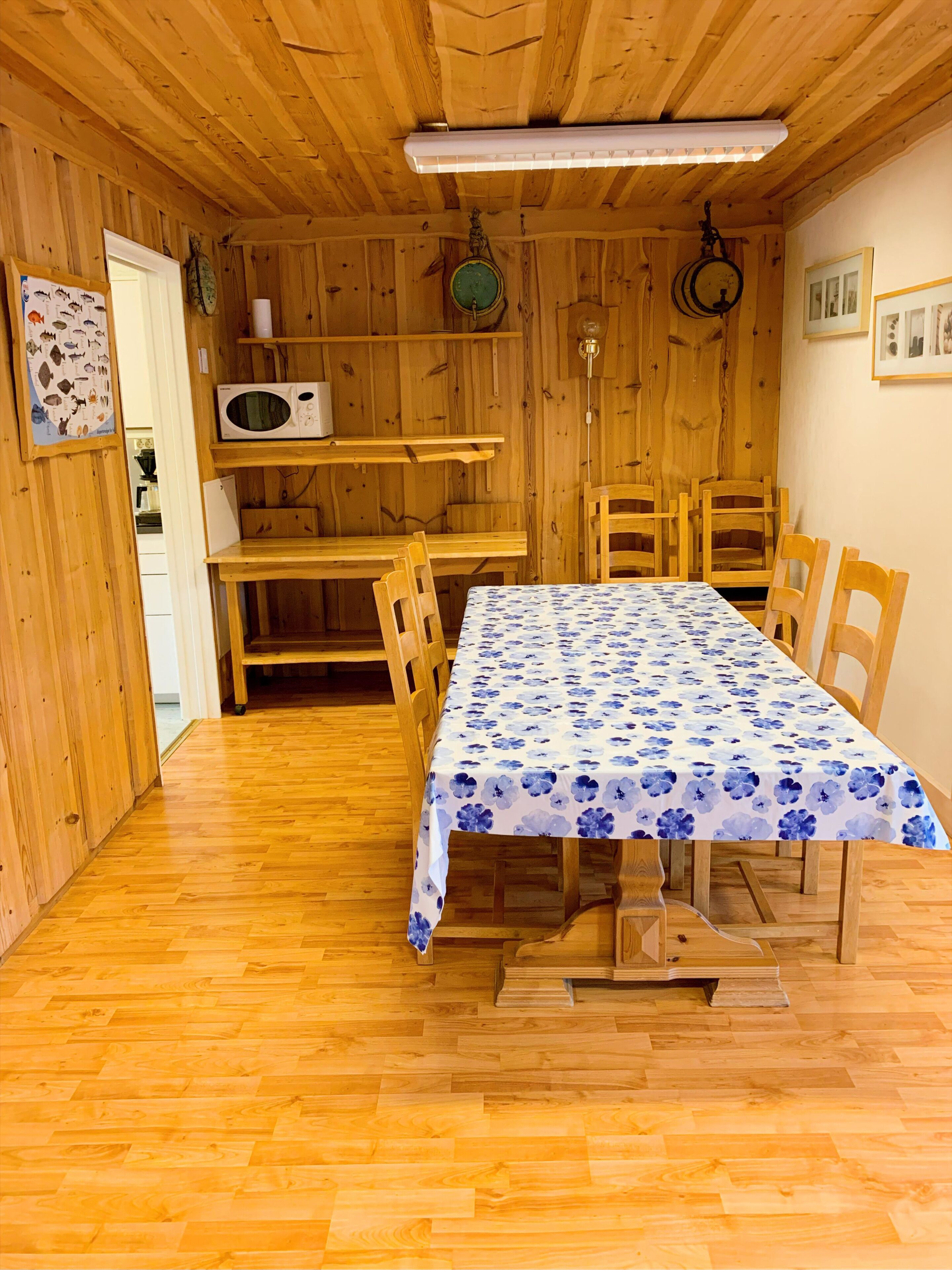 A wooden interior with a dining table covered by a floral tablecloth, wooden chairs, a microwave, and a clock on the wall.