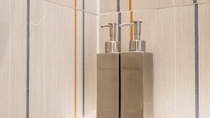 To metal dispensere til toiletartikler, mærket 'Hotel Olive Shampoo' og 'Skyl', står på en hvid hylde foran en stribet væg.