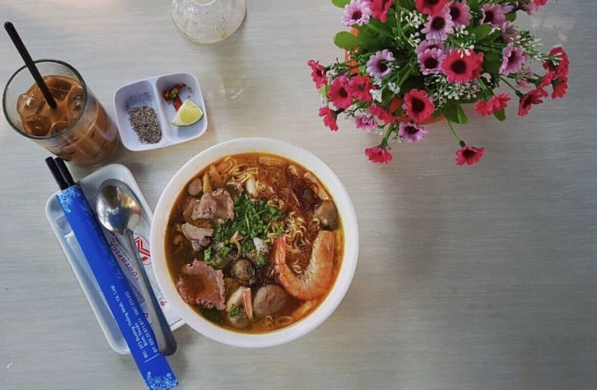 Kulhollinen keittoa, jossa on erilaisia aineksia, lasillinen kylmää juomaa, lusikka, pieni kulho mausteita ja maljakko kukkia pöydällä.