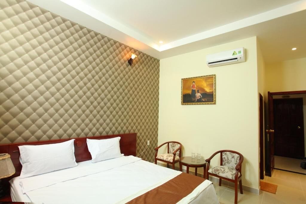 Habitación de hotel con una cama grande, dos sillas, una mesita pequeña, un equipo de aire acondicionado y un cuadro en la pared.