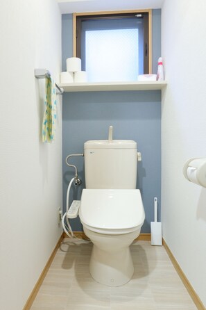 A small bathroom with a toilet, a shelf with toilet paper, a window, and a towel hanging on the wall. - Cross Stage (Fukuoka)