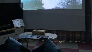 Huoneessa on suuri näyttö, jossa esitetään merimaisema, litteänäyttöinen televisio, pieni pöytä, jossa on kaukosäätimet, ja sohva.