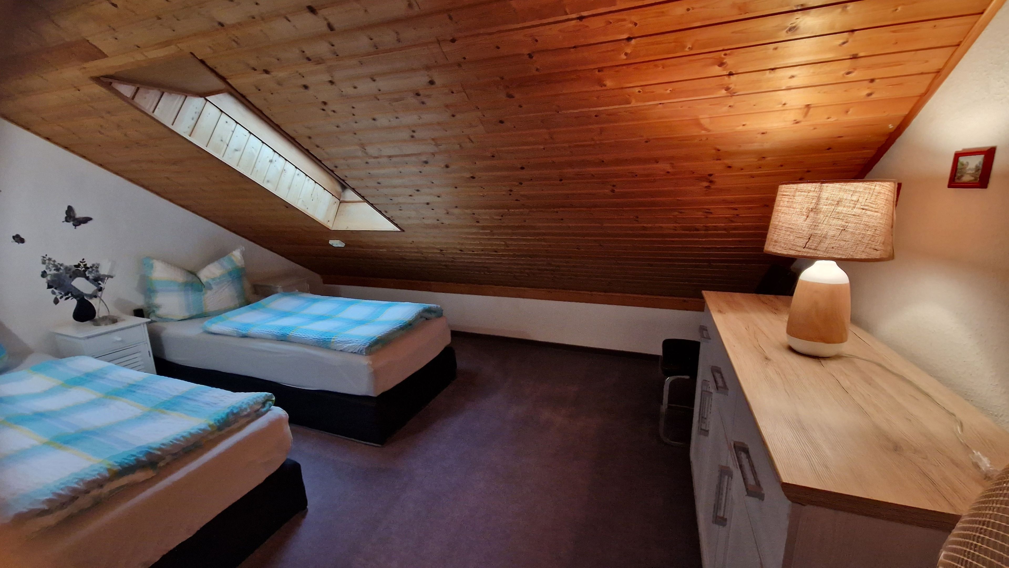Ein Schlafzimmer mit zwei Betten, einem Oberlicht, einer Holzbalkendecke, einem Nachttisch mit Lampe und einer kleinen Kommode.